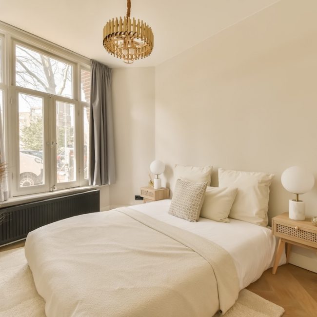 a bedroom with white walls and hardwood flooring the room has a large bed, a wooden side table and two windows