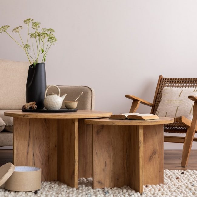 Interior design of living room with beige sofa, rattan armchair, coffee tables, tea pot, flowers in vase, ladder, rug and elegant decoration. Beige wall. Cozy home decor. Template.