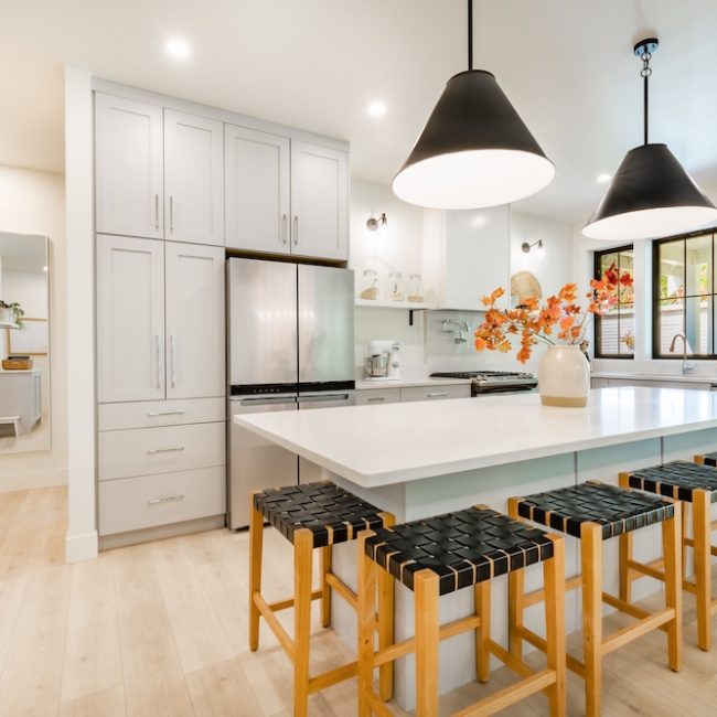 Modern farmhouse kitchen interior with light wood floors white granite marble counters large dining table with eight chairs stainless appliances orange color accents and large windows with black trim