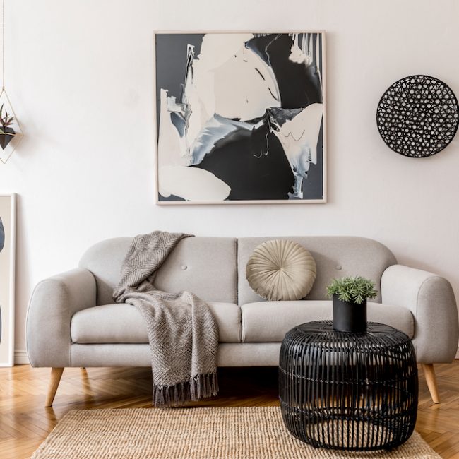 Stylish and scandinavian living room interior of modern apartment with gray sofa, pillows, plaid, plants, design wooden commode, black table, lamp, abstrac paintings on the wall.  Modern home decor.